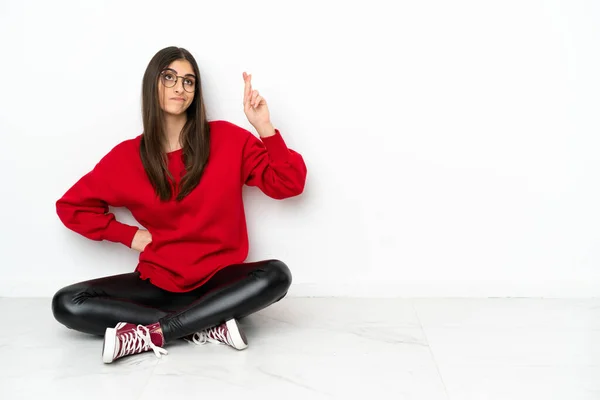 Young woman sitting on the floor isolated on white background with fingers crossing and wishing the best