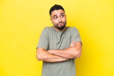 Genç Arap yakışıklı adam sarı arka planda izole edilmiş. Omuzlarını kaldırırken şüpheyle hareket ediyor.
