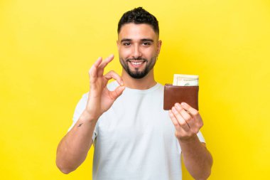 Sarı arka planda izole edilmiş cüzdan taşıyan genç bir Arap, tamam işaretini parmaklarıyla gösteriyor.