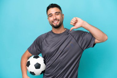 Genç Arap yakışıklı adam mavi arka planda izole edilmiş futbol topuyla ve kendisiyle gurur duyuyor.