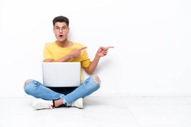 Genç adam beyaz arka planda izole bir şekilde yerde oturuyor. Şaşırmış ve işaret ediyor.