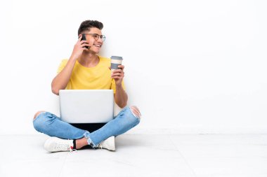 Genç adam beyaz arka planda izole bir şekilde yerde oturuyor. Elinde kahve ve cep telefonu var.