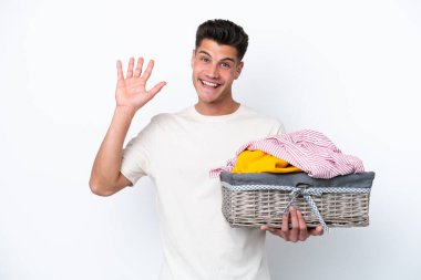 Çamaşır sepetini tutan beyaz tenli genç adam beyaz arka planda mutlu bir ifadeyle selam veriyor.