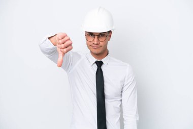 Young architect caucasian man with helmet and holding blueprints isolated on white background showing thumb down with negative expression
