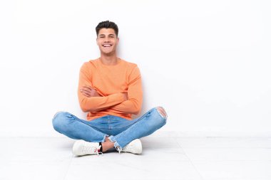 Young man sitting on the floor isolated on white background keeping the arms crossed in frontal position