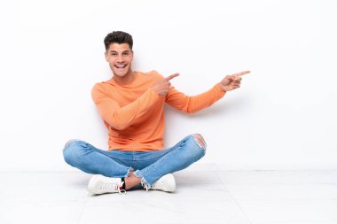 Genç adam, beyaz arka planda izole bir şekilde yerde oturuyor. Parmağını kenarı gösteriyor ve bir ürün sunuyor.