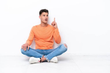 Young man sitting on the floor isolated on white background thinking an idea pointing the finger up