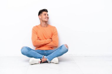 Young man sitting on the floor isolated on white background looking up while smiling
