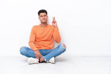 Young man sitting on the floor isolated on white background with fingers crossing and wishing the best