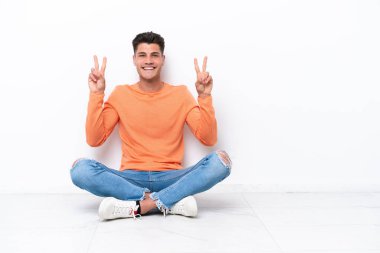Young man sitting on the floor isolated on white background showing victory sign with both hands