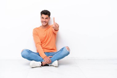 Young man sitting on the floor isolated on white background showing and lifting a finger