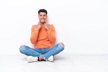 Young man sitting on the floor isolated on white background smiling with a happy and pleasant expression
