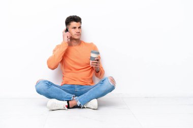 Genç adam beyaz arka planda izole bir şekilde yerde oturuyor. Elinde kahve ve cep telefonu var.