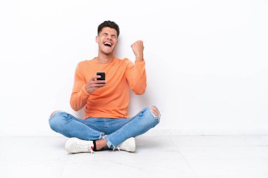 Young man sitting on the floor isolated on white background with phone in victory position