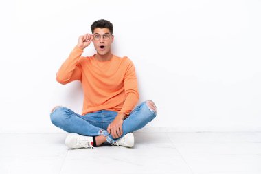 Genç adam beyaz arka planda gözlüklü ve şaşırmış bir şekilde yerde oturuyor.