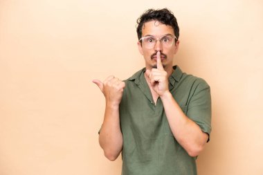 Beyaz tenli genç adam bej arka planda izole edilmiş. Yan tarafı işaret ediyor ve sessizlik hareketi yapıyor.