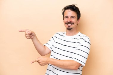 Beyaz tenli genç adam bej arka planda izole edildi. Parmağını yana uzattı ve bir ürün sundu.