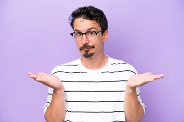 Young caucasian man isolated on purple background With glasses and having doubts