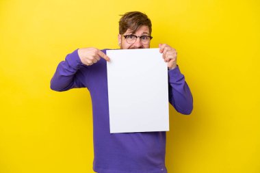 Sarı arka planda sakallı kızıl saçlı adam boş bir levha tutuyor ve arkasına saklanıyor.