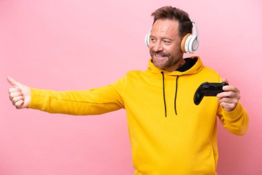 Orta yaşlı bir adam pembe arka planda izole edilmiş bir bilgisayar oyunu kontrolörüyle oynarken başparmak hareketi yapıyor.