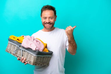 Orta yaşlı bir kadın mavi arka planda izole edilmiş bir çamaşır sepetini elinde tutuyor. Ürünü sunmak için yan tarafa bakıyor.