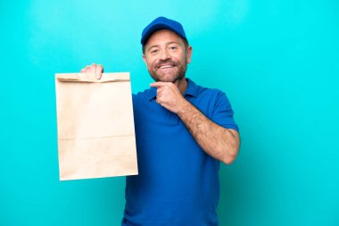 Orta yaşlı bir adam mavi arka planda izole edilmiş bir paket yiyecek alıyor mutlu ve gülümsüyor.