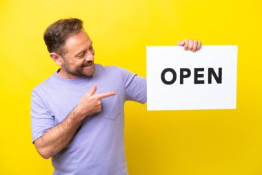 Orta yaşlı, beyaz bir adam sarı arka planda izole edilmiş. Elinde açık yazılı bir pankart var.