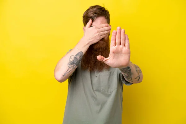Redhead man with beard isolated on yellow background making stop gesture and covering face