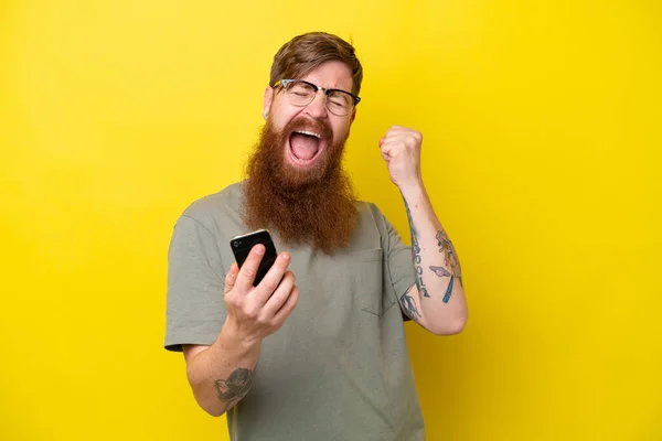 Rudowłosy Mężczyzna Brodą Odizolowany Żółtym Tle Telefonem Pozycji Zwycięstwa — Zdjęcie stockowe