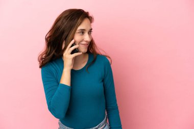 Pembe arka planda izole edilmiş genç beyaz bir kadın cep telefonuyla sohbet ediyor.