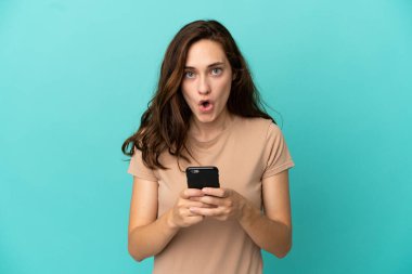 Mavi arka planda izole edilmiş genç beyaz kadın kameraya bakarken şaşırmış bir ifadeyle cep telefonunu kullanıyor.