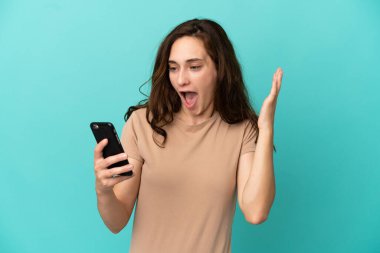 Mavi arka planda izole edilmiş genç beyaz kadın kameraya bakarken şaşırmış bir ifadeyle cep telefonunu kullanıyor.