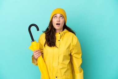 Mavi arka planda şemsiye tutan beyaz bir adam yukarı bakıyor ve şaşırmış bir ifadeyle