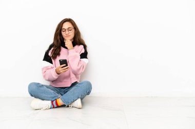 Beyaz arka planda izole bir şekilde yerde oturan beyaz bir kadın düşünüyor ve bir mesaj gönderiyor.