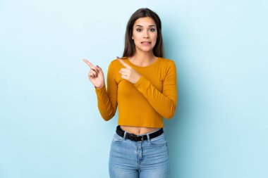 Mavi arka planda izole edilmiş genç beyaz kadın korkmuş ve kenarı işaret ediyor.