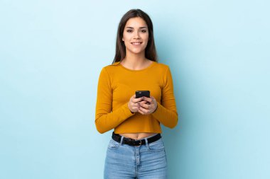 Mavi arka planda izole edilmiş genç beyaz kadın cep telefonuyla bir mesaj gönderiyor.