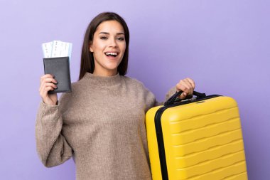 Mor arka planda izole edilmiş genç beyaz bir kadın, bavul ve pasaportla tatilde.