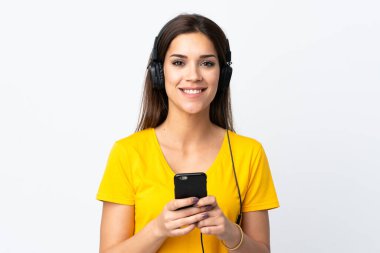 Beyaz arka planda izole edilmiş genç beyaz bir kadın cep telefonuyla müzik dinliyor ve ön tarafa bakıyor.