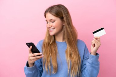Pembe arka planda izole edilmiş genç sarışın kadın kredi kartıyla cep telefonuyla alışveriş yapıyor.