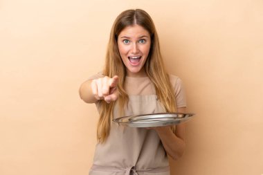 Bej arka planda saklanmış tepsisi olan genç sarışın garson kadın şaşırmış ve ön tarafı işaret ediyor.