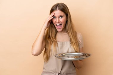 Tepsisi bej arka planda, sürpriz ifadeli genç sarışın garson kadın.