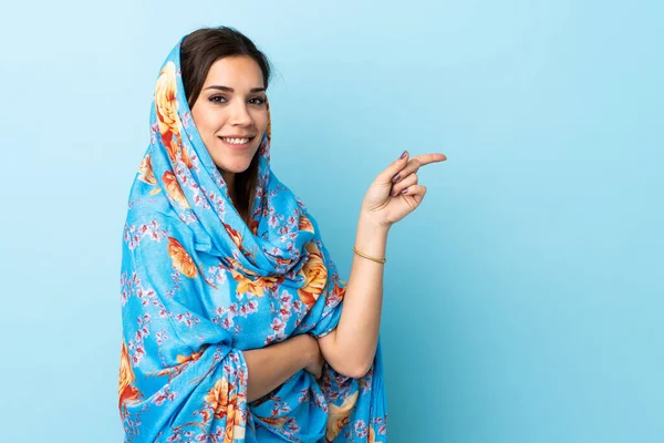 伝統的な衣装を身に着けている若いモロッコの女性は 側に指を指す青の背景に隔離された — ストック写真