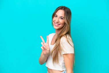 Mavi arka planda izole edilmiş genç beyaz kadın gülümsüyor ve zafer işareti gösteriyor.