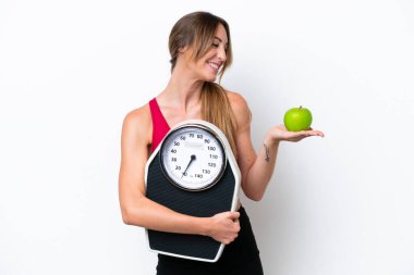 Beyaz arka planda izole edilmiş genç beyaz bir kadın elmaya bakarken bir tartı makinesi tutuyor.