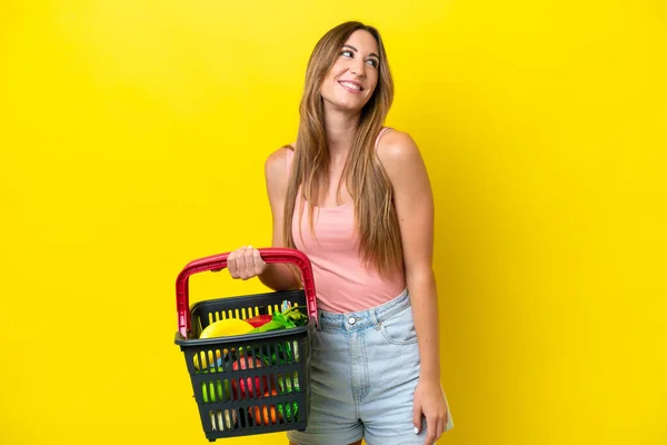 黄色の背景に孤立した食べ物でいっぱいの買い物かごを持っている若い白人女性は アイデアを考えながら検索します — ストック写真