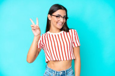Mavi arka planda izole edilmiş genç beyaz kadın gülümsüyor ve zafer işareti gösteriyor.