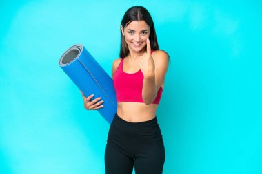 Genç beyaz kadın yoga derslerine giderken mavi arka planda izole edilmiş bir minder tutuyor.