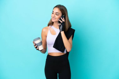 Mavi arka planda izole edilmiş genç bir kadın elinde kahve ve bir cep telefonu tutuyor.