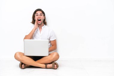 Yerde dizüstü bilgisayarı olan yakışıklı genç bir adam beyaz arka planda izole edilmiş, şaşırmış ve şok olmuş bir yüz ifadesi var.