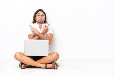 Yerde dizüstü bilgisayarı olan genç ve yakışıklı bir adam beyaz arka planda izole bir şekilde oturuyor.